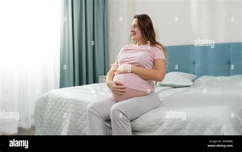 Beautiful Smiling Pregnant Woman In Pajamas Sitting On Bed And Stroking