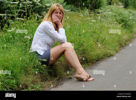 Mature Woman Wearing A White Shirt And Skirt Sitting On The Roadside