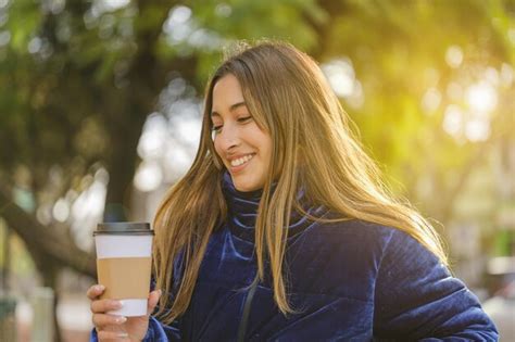 Hermosa chica latina sonriente bebiendo café en un banco de un parque público Foto Premium