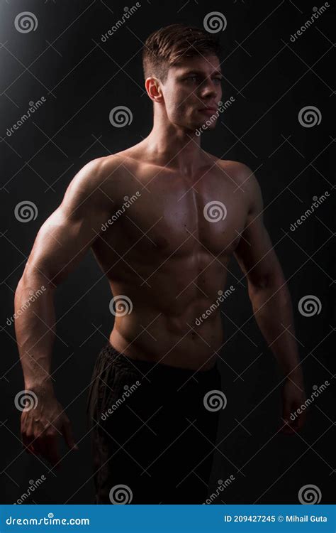 Muscular Bodybuilder Posing With A Naked Torso On A Black Background In The Dark With Backlight