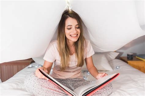Souriante Blonde Jeune Femme Assise Sur Le Lit En Lisant Le Magazine Photo Gratuite