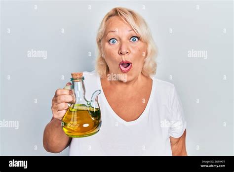 Middle Age Blonde Woman Holding Olive Oil Can Scared And Amazed With Open Mouth For Surprise