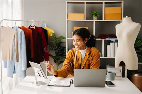 Calm Curly Brunette Dark Skinned Woman On Desk In Office Of Fashion Designer And Holds Tablet
