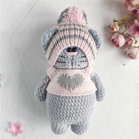 A Crocheted Teddy Bear With A Pink Hat And Scarf Sitting Next To Flowers