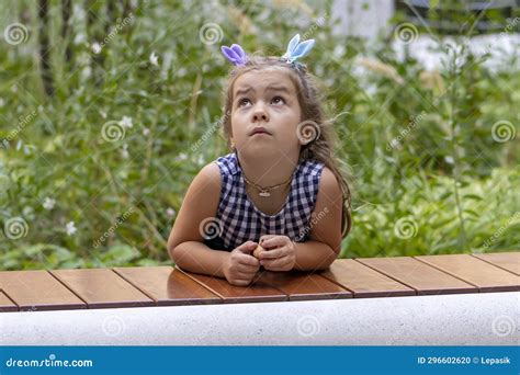 A Blonde Year Old Girl Looks Up Pensively Against A Backdrop Of Greenery And Nature Stock