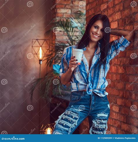 A Brunette In A Flannel Shirt And Jeans Holds A Cup Of Coffee Posing Against A Brick Wall Stock