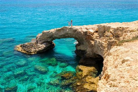 two people are standing on the edge of a rock arch over clear blue ...