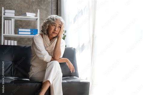 Depressed Asian Senior Woman Sit On Sofa Looking Away By The Window