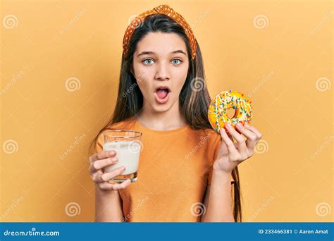 Young Brunette Girl Eating Donut And Drinking Glass Of Milk Afraid And