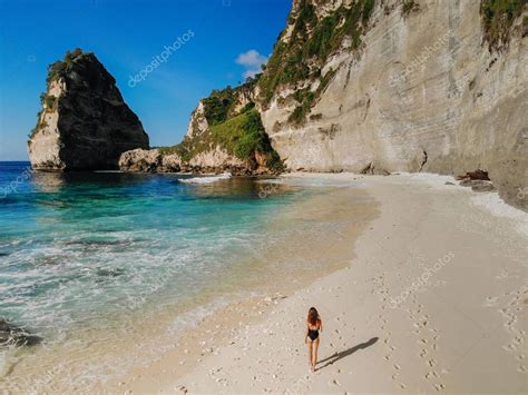 Mujer en bikini negro caminando por la playa con rocas marinas y océano turquesa cielo azul