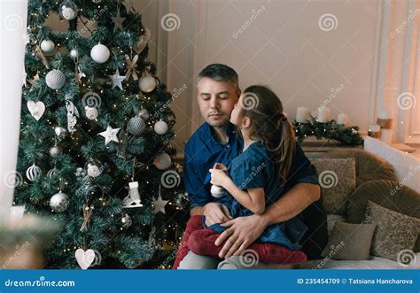 A Little Daughter Gently Kisses Her Dad On The Cheek While Decorating A Christmas Tree Waiting