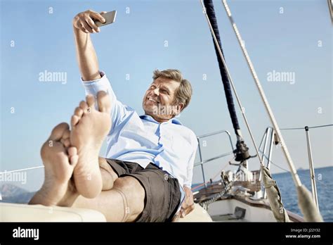 Smiling Mature Man Lying On Deck Of His Sailing Boat Taking Selfie With Cell Phone Stock Photo