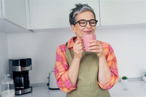 Charming Mature Woman In Apron Enjoys A Warm Drink In Her Cozy Kitchen Embracing Leisure And