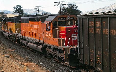 an orange and black train traveling down tracks next to mountains with ...