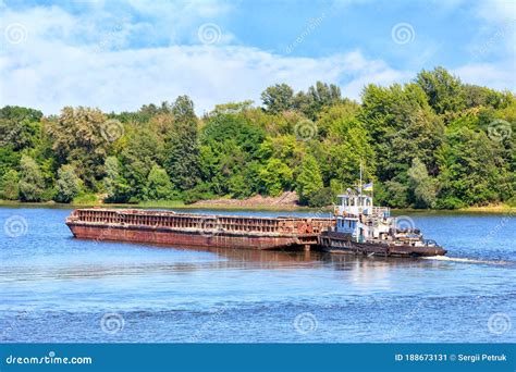 A River Tug Is Pushing A Rusty Barge Along The River Along The Shore