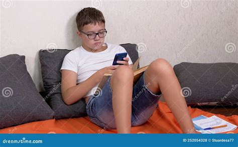 Teen In Glasses With A Smartphone In Hands And Open Book Studying