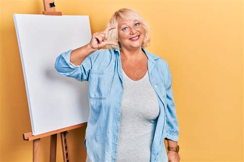 Middle Age Blonde Woman Standing By Painter Easel Stand Smiling And