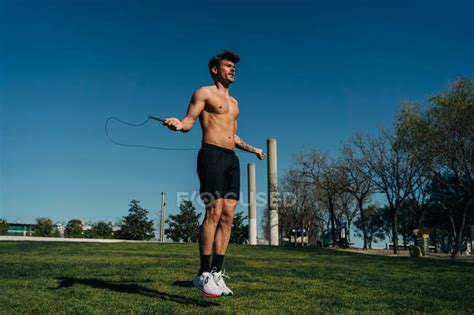Male Athlete In Naked Torso Jumping With Skipping Rope And Looking Away On Walkway During Cardio