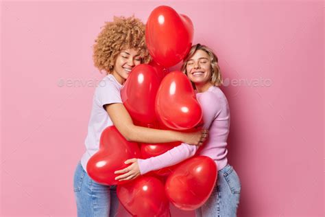 Two Positive Young Women Embrace Bunch Of Inflated Red Heart Shaped