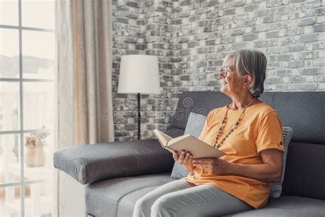 Happy Older Blonde Woman Sitting On Comfortable Sofa In Living Room Reading Interesting Book