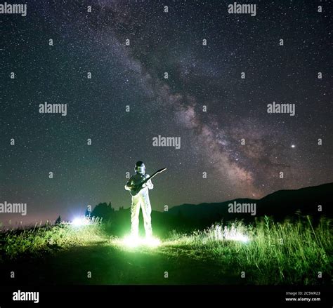 Spaceman in space suit playing guitar in mountain valley under magical night sky with Milky Way ...