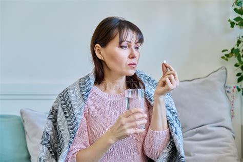 Sick Mature Woman Taking Pill With Glass Of Water Sitting Under Blanket At Home Stock Photo