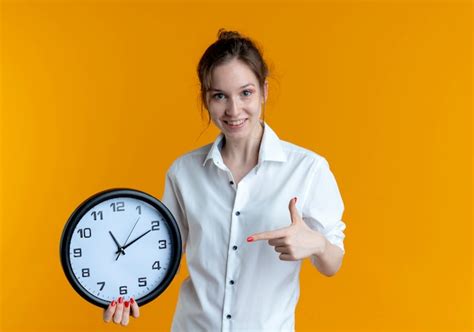 Jeune fille russe blonde souriante tient et pointe à l horloge isolée sur l espace orange avec