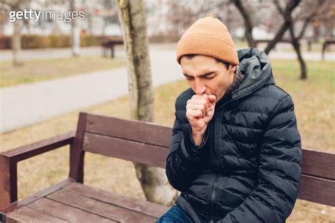 Young Sick Man Coughing Sitting On Bench In Fall Park Covering Mouth