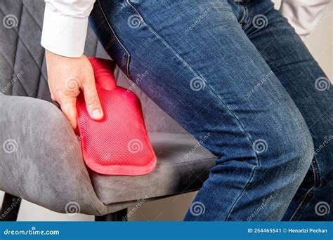 A Male Office Worker Sits On A Chair And Fixes A Red Heating Pad With Hot Water The Concept Of