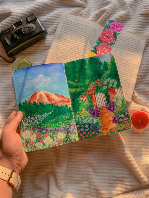 A Person Holding An Open Book On Top Of A Bed Next To Flowers And A Camera