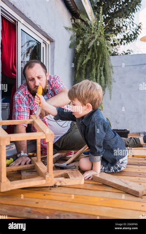 Mature Dad Explaining To Son How To Drive Screw Into Wooden Block While Sitting On Boardwalk