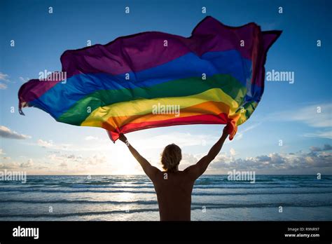 Silhouette Of Man Holding A Gay Pride Rainbow Flag Fluttering Against The Rising Sun On An Empty