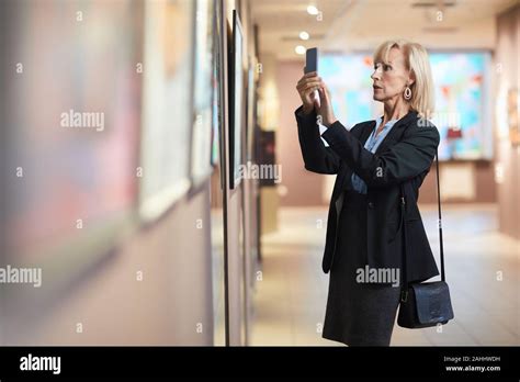 Side View Portrait Of Elegant Mature Woman Taking Smartphone Photo Of Painting While Enjoying