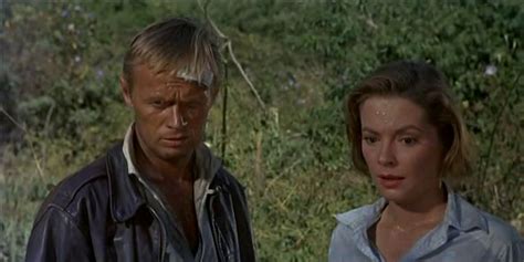 A Man And Woman Standing Next To Each Other In Front Of Some Bushes