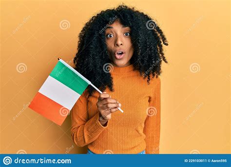 African American Woman With Afro Hair Holding Ireland Flag Scared And Amazed With Open Mouth For