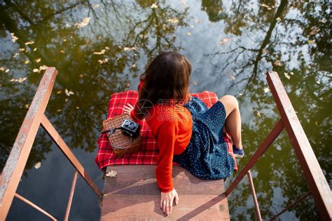 男人一位年轻女子在阳光明媚的日子里背坐在湖边的一座桥上格子布相机手提箱一位年轻女子在阳光明媚的日子里背坐在湖边的一座桥上格子布手高清图片下载