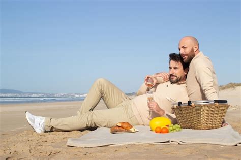 Hombre calvo bebiendo champán con su novio en un picnic en la playa guapa pareja gay barbuda