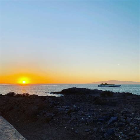 the sun is setting over the ocean with rocks on the shore and a boat in ...