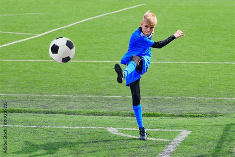 Teenage Blonde Caucasian Soccer Football Player In Blue Sport Uniform Is Kicking Ball In