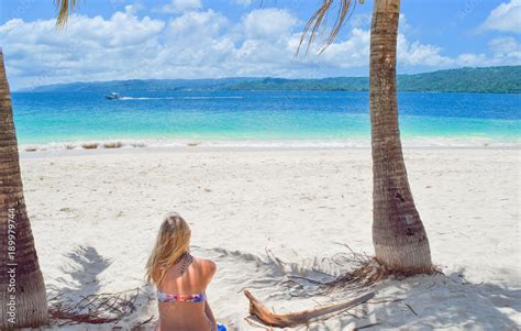Frau sitzt im Bikini auf Handtuch mit dem Rücken zur Kamera am weißen Sandstrand und schaut auf