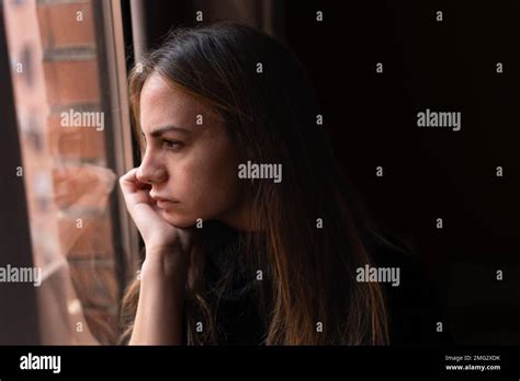 Side view of young thoughtful female with long dark hair leaning chin on hand and looking out ...