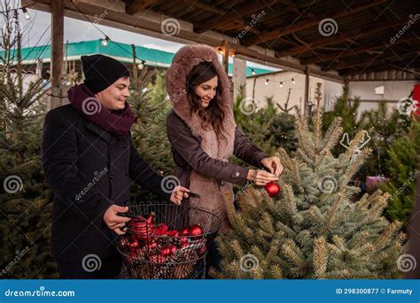 Funny Couple Decorates Christmas Trees At Market With Red Balls Man And Woman Hang Toys On Fir