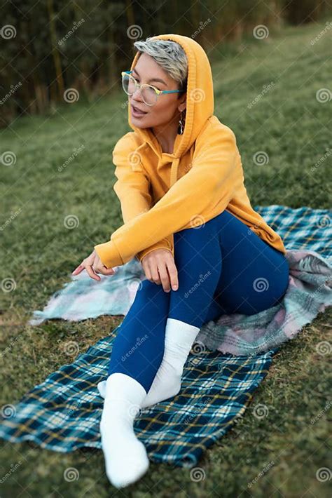 Cute Girl With Glasses In An Orange Hoodie Is Sitting In A Park In