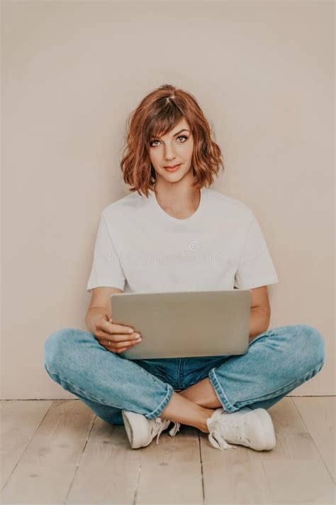 A Brunette Sits On The Floor With A Laptop On A Beige Wall Background She Is Wearing A White T