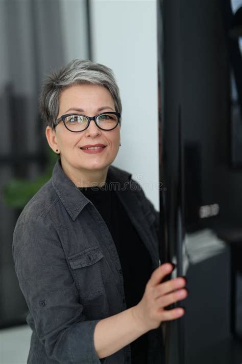 Positive Mature Woman Opens The Refrigerator In The Kitchen Vertical Portrait Stock Photo
