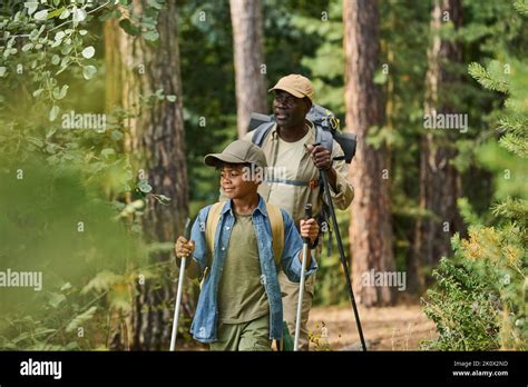 Mature Black Man And His Grandson With Trekking Sticks Moving Down Footpath Along Green Trees