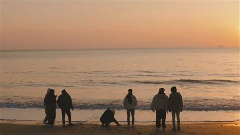 A Group Of People Standing On Top Of A Beach Next To The Ocean At Sunset