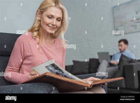 Selective Focus Of Smiling Mature Woman With Photo Album And Son Using Laptop On Sofa At Home