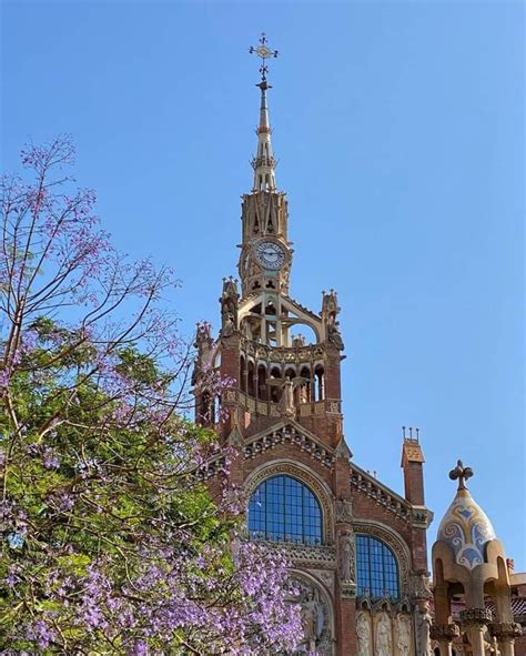 A Tall Building With A Clock On It S Side And Purple Flowers In The