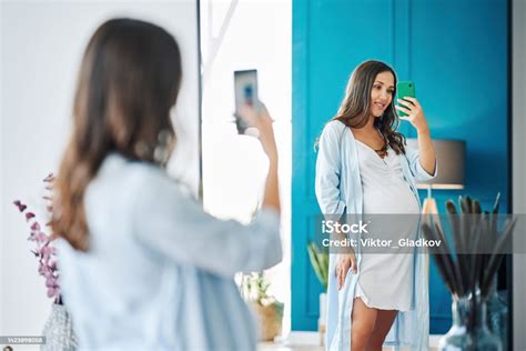 Portrait Of Smiling Pregnant Woman Taking Mirror Selfie At Modern Home